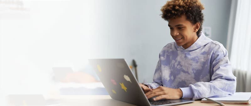 Smiling student in a tie-dye hoodie using a laptop with stickers, studying in a bright room